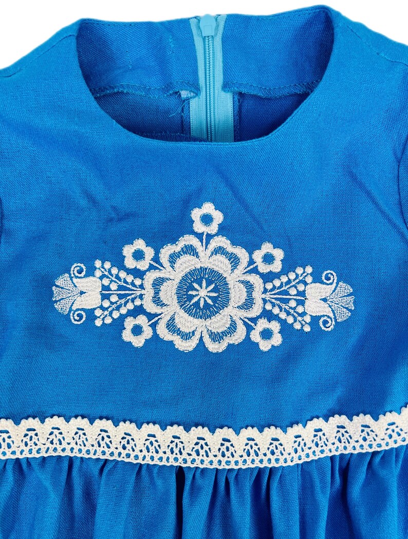 K&ouml;nnte beinhalten: Ein blaues Leinenkleid mit wei&szlig;en gestickten Blumenmotiven und einem wei&szlig;en Spitzenbesatz am Saum.