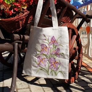 Op de afbeelding: Een witte linnen tas met geborduurde paarse irissen. De tas hangt aan een houten kar met een rood wiel.