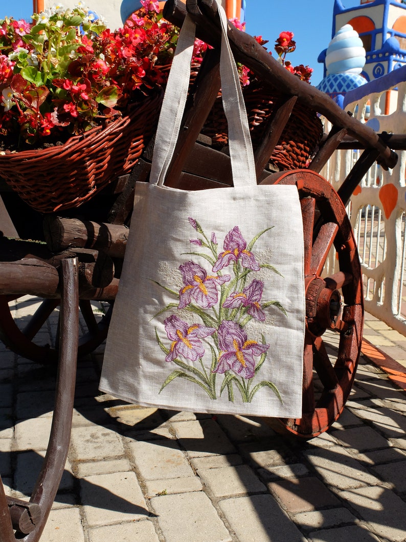 Op de afbeelding: Een witte linnen tas met geborduurde paarse irissen. De tas hangt aan een houten kar met een rood wiel.
