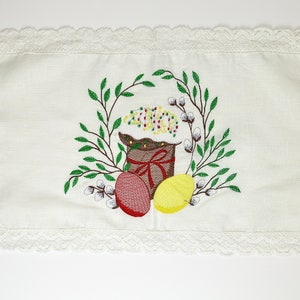 May include: White rectangular Easter table runner with a delicate lace trim. The center features an embroidered design of an Easter cake, eggs, and a wreath of green leaves and pussy willows.