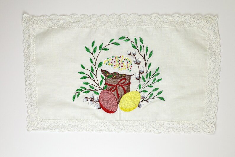 May include: White rectangular Easter table runner with a delicate lace trim. The center features an embroidered design of an Easter cake, eggs, and a wreath of green leaves and pussy willows.