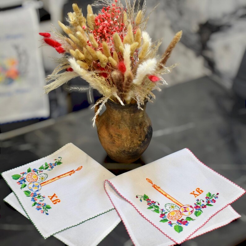 Puede incluir: Dos servilletas de lino bordadas blancas con un borde verde y rojo. Las servilletas presentan una vela, un huevo y el texto "XB". Las servilletas est&aacute;n sobre una mesa con un jarr&oacute;n de flores secas.
