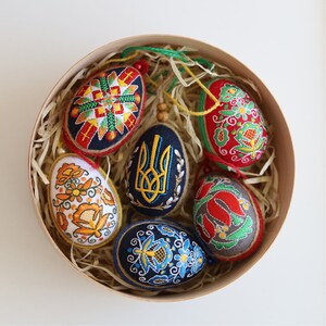 K&ouml;nnte beinhalten: Sechs bestickte Ostereier in einer runden Holzkiste. Die Eier sind mit traditionellen ukrainischen Volksmotiven verziert, darunter ein gelbes und blaues Dreizack, ein Blumenmuster und ein geometrisches Muster. Die Eier liegen in zerkn&uuml;lltem Papier.