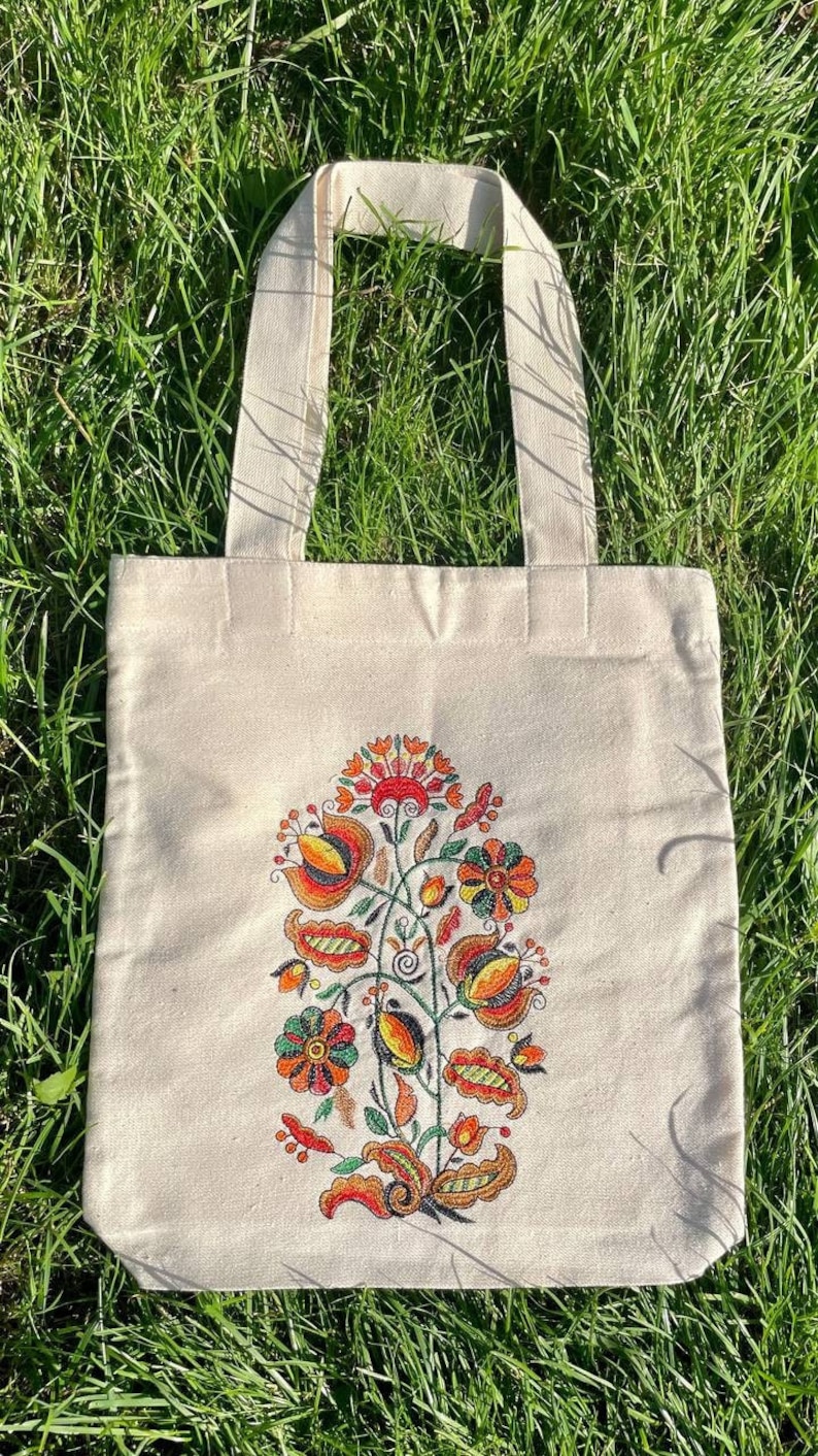 May include: A white canvas tote bag with a floral design embroidered in orange, red, yellow, and green. The design features a stylized flower with leaves and stems.