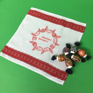May include: White embroidered kitchen towel with red trim and the text "Хриcтоc Воскрес!" (Christ is Risen!) in a wreath of stylized eggs. The towel is decorated with a red and black embroidered border.  The towel is lying on a green surface with several colorful, hand-painted Easter eggs.