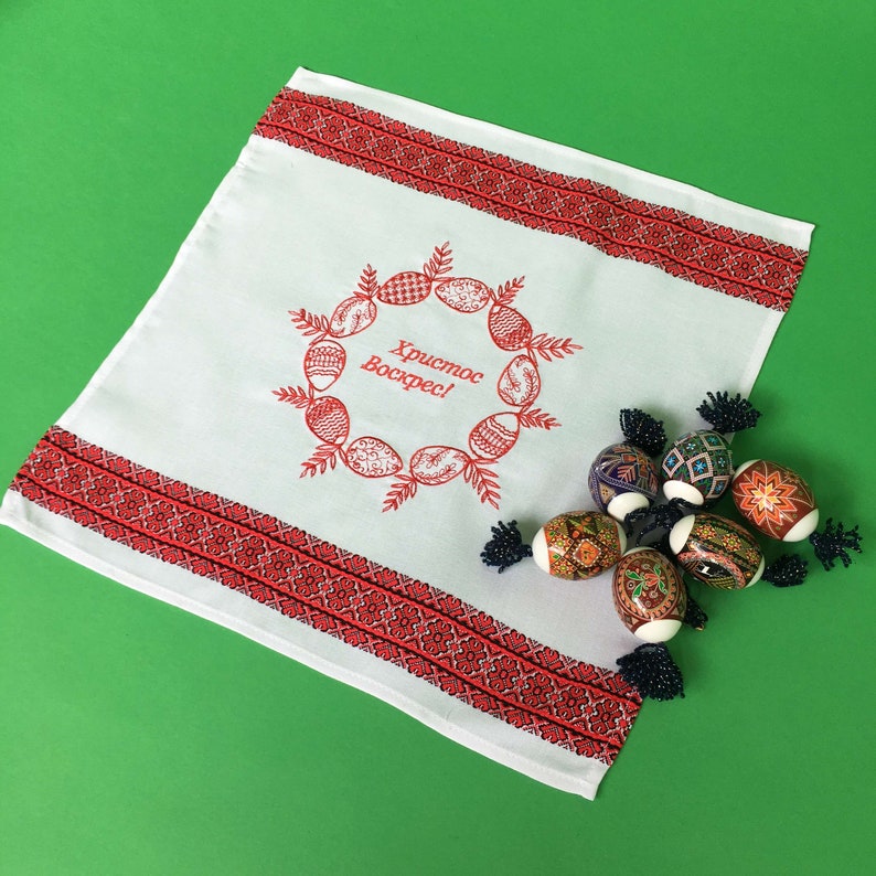 May include: White embroidered kitchen towel with red trim and the text "Хриcтоc Воскрес!" (Christ is Risen!) in a wreath of stylized eggs. The towel is decorated with a red and black embroidered border.  The towel is lying on a green surface with several colorful, hand-painted Easter eggs.