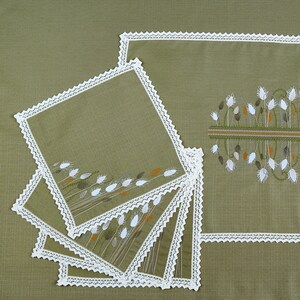 May include: A set of embroidered placemats and matching coasters. The placemats and coasters are olive green with white lace trim. The embroidery features wheat stalks in white, brown, and orange. The placemats are rectangular, and the coasters are square.