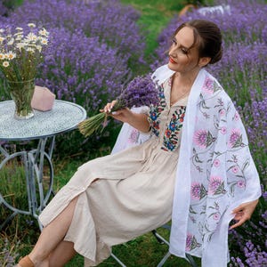 May include: A woman in a beige dress and white shawl with a floral print sits in a lavender field. She holds a bouquet of lavender. A small metal table with a vase of white flowers and a pink face mask is nearby.