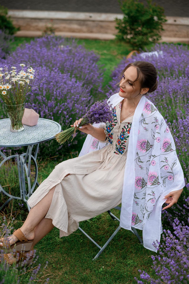 May include: A woman in a beige dress and white shawl with a floral print sits in a lavender field. She holds a bouquet of lavender. A small metal table with a vase of white flowers and a pink face mask is nearby.