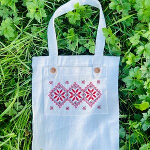 Op de afbeelding: Een witte linnen tote bag met een rood en zwart geborduurd ontwerp. De tas heeft twee bruine knopen aan de voorkant.