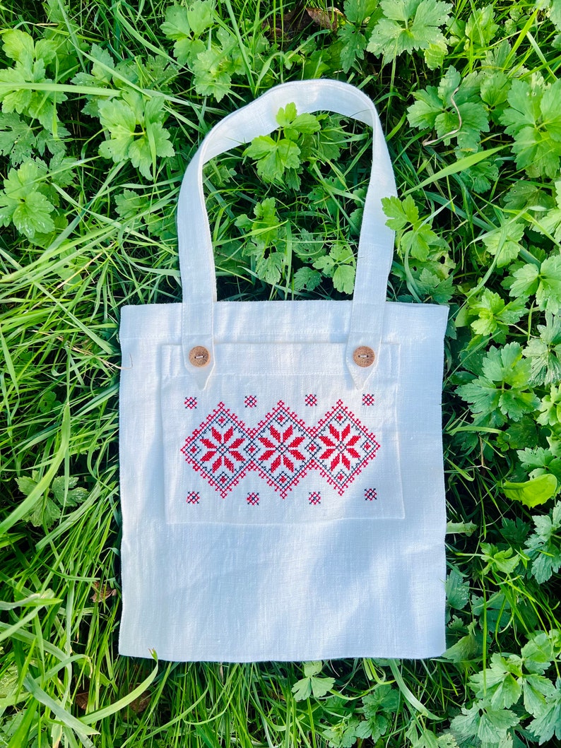 Op de afbeelding: Een witte linnen tote bag met een rood en zwart geborduurd ontwerp. De tas heeft twee bruine knopen aan de voorkant.