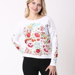 Puede incluir: Camiseta blanca de manga larga con un dise&ntilde;o bordado de flores y p&aacute;jaros coloridos.