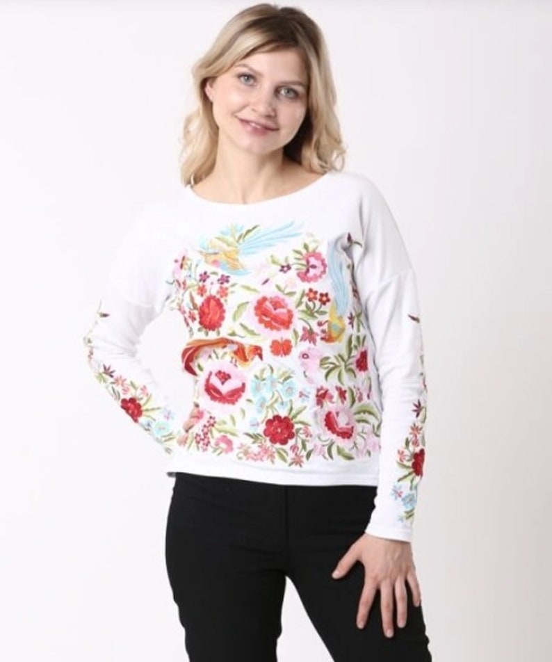 Puede incluir: Camiseta blanca de manga larga con un dise&ntilde;o bordado de flores y p&aacute;jaros coloridos.
