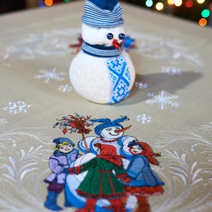 Op de afbeelding: Een handgemaakte sneeuwpop van witte stof, met een blauw gestreepte hoed, sjaal en decoratieve patch. De sneeuwpop heeft knoopogen en een rode neus. Tafelkleed met een winterse sc&egrave;ne met kinderen en een sneeuwpop.