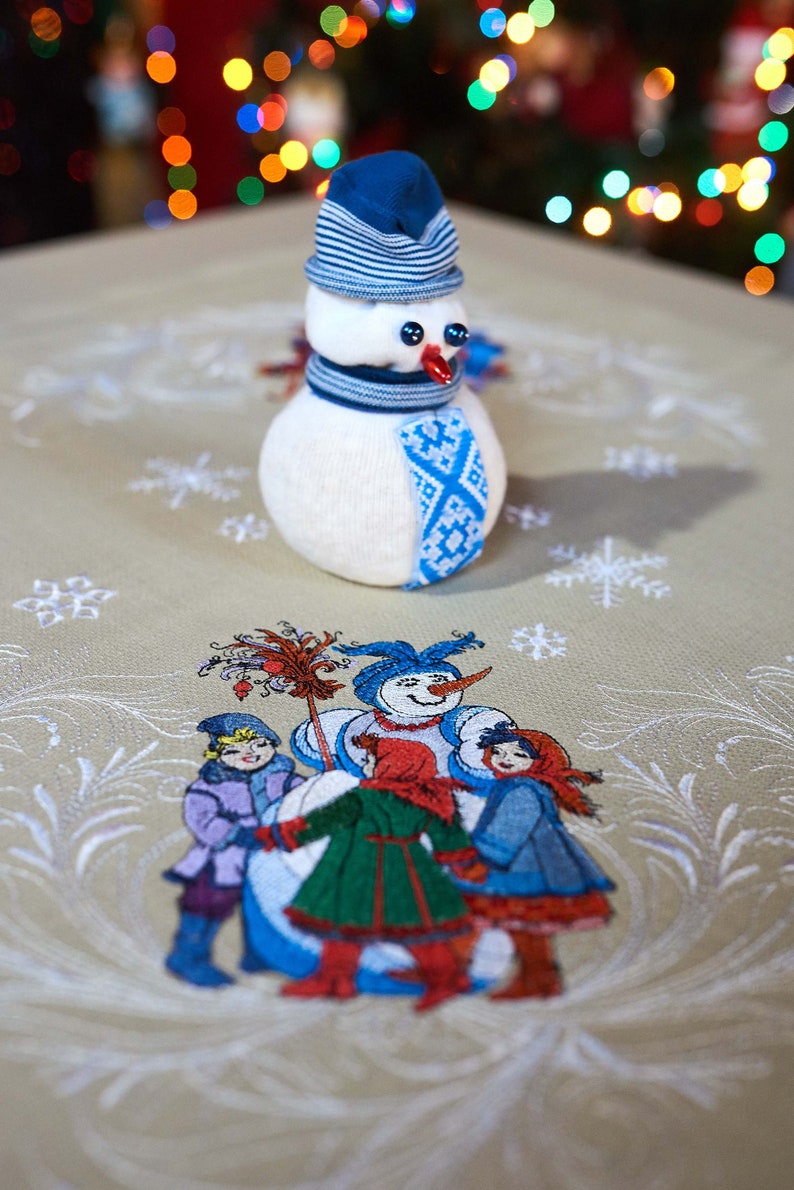 Op de afbeelding: Een handgemaakte sneeuwpop van witte stof, met een blauw gestreepte hoed, sjaal en decoratieve patch. De sneeuwpop heeft knoopogen en een rode neus. Tafelkleed met een winterse sc&egrave;ne met kinderen en een sneeuwpop.