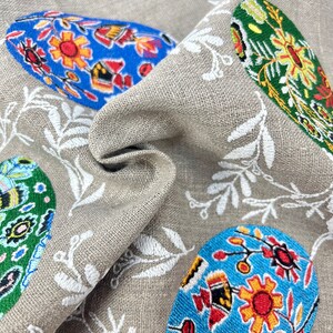 以下が含まれることがあります： ベージュのリネン生地に、白い刺繍の花柄とカラフルなイースターエッグが施されています。卵は、青、緑、赤、黄、黒の花柄で飾られています。