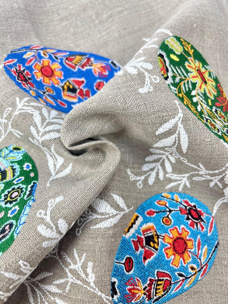以下が含まれることがあります： ベージュのリネン生地に、白い刺繍の花柄とカラフルなイースターエッグが施されています。卵は、青、緑、赤、黄、黒の花柄で飾られています。