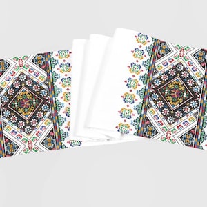 May include: Two white table runners with embroidered geometric patterns in black, red, yellow, green, and blue. The patterns feature a central diamond shape with smaller floral motifs.