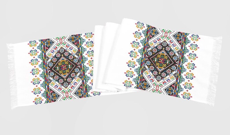 May include: Two white table runners with embroidered geometric patterns in black, red, yellow, green, and blue. The patterns feature a central diamond shape with smaller floral motifs.