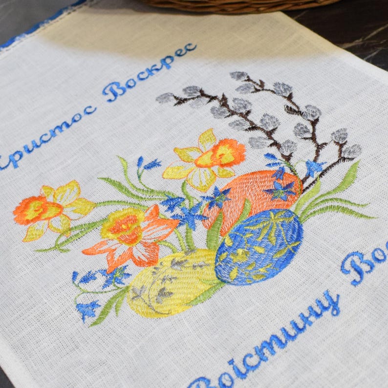 Puede incluir: Toalla de cocina blanca bordada con un dise&ntilde;o floral de narcisos amarillos, flores azules y ramas de sauce grises. La toalla tambi&eacute;n presenta dos frases bordadas en azul: "Christos Voskres" y "Voistinu Voskres".