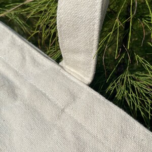May include: Close-up of a white canvas tote bag with a thick white canvas strap. The strap is sewn to the bag with white thread.