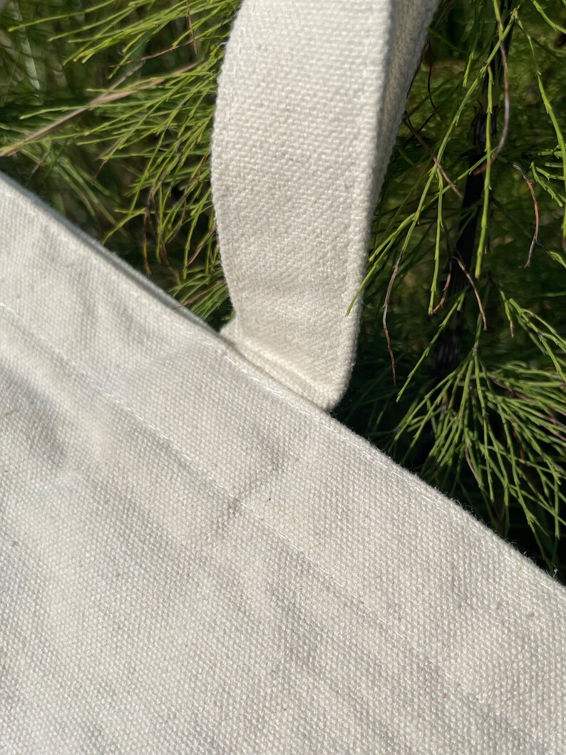 May include: Close-up of a white canvas tote bag with a thick white canvas strap. The strap is sewn to the bag with white thread.