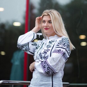 Puede incluir: Blusa blanca con mangas largas y bordados florales negros intrincados. La blusa presenta un cuello alto y se combina con pantalones negros. Las mangas tienen un patr&oacute;n de encaje y el estilo es tradicional.