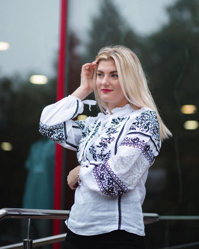 Puede incluir: Blusa blanca con mangas largas y bordados florales negros intrincados. La blusa presenta un cuello alto y se combina con pantalones negros. Las mangas tienen un patr&oacute;n de encaje y el estilo es tradicional.