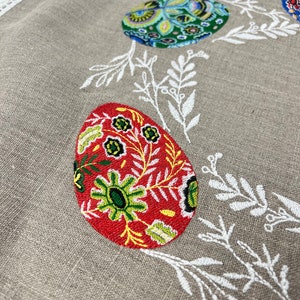 以下が含まれることがあります： ベージュのリネン製のテーブルクロスに、白い刺繍の花柄とカラフルな刺繍の卵が施されています。卵は、赤、緑、青、黄色の複雑な模様で飾られています。