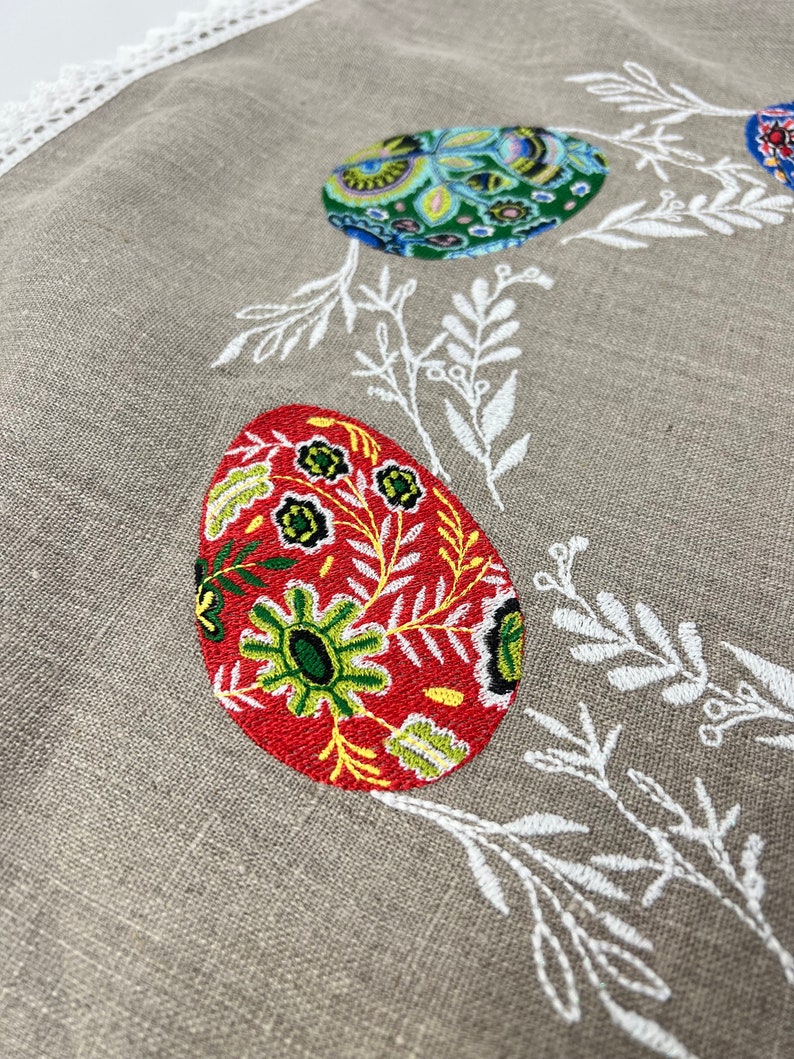 以下が含まれることがあります： ベージュのリネン製のテーブルクロスに、白い刺繍の花柄とカラフルな刺繍の卵が施されています。卵は、赤、緑、青、黄色の複雑な模様で飾られています。