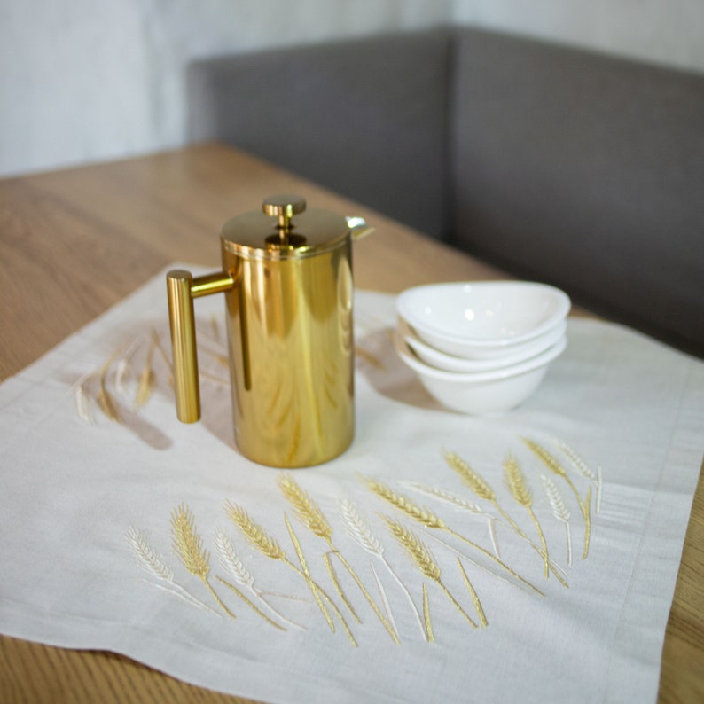 Peut inclure: Une cafeti&egrave;re &agrave; piston dor&eacute;e avec une poign&eacute;e et un couvercle, pos&eacute;e sur un tissu en lin blanc brod&eacute; d'&eacute;pis de bl&eacute;. Trois petits bols blancs sont empil&eacute;s &agrave; proximit&eacute;. La sc&egrave;ne se d&eacute;roule sur une table en bois.