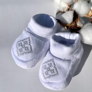 K&ouml;nnte beinhalten: Ein Paar wei&szlig;e Baby-Booties mit silbernem Stickdesign auf der Vorderseite. Die Booties haben eine weiche, pl&uuml;schige Textur und einen gerollten Bund. Baumwollbl&uuml;ten sind im Hintergrund.