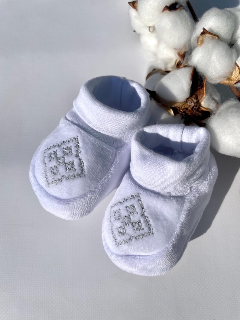 K&ouml;nnte beinhalten: Ein Paar wei&szlig;e Baby-Booties mit silbernem Stickdesign auf der Vorderseite. Die Booties haben eine weiche, pl&uuml;schige Textur und einen gerollten Bund. Baumwollbl&uuml;ten sind im Hintergrund.