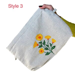 May include: A beige canvas zippered pouch with a floral embroidery design. The embroidery features yellow flowers with orange centers and green leaves.