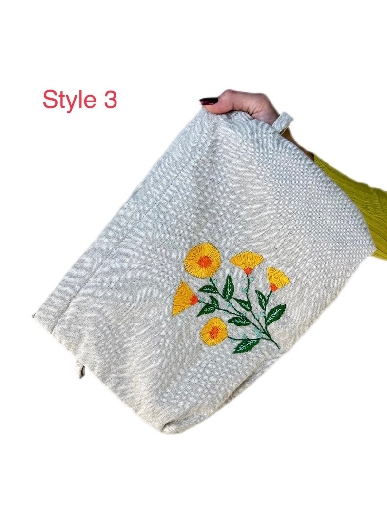 May include: A beige canvas zippered pouch with a floral embroidery design. The embroidery features yellow flowers with orange centers and green leaves.