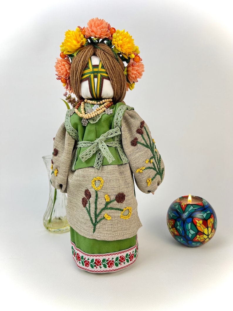Puede incluir: Mu&ntilde;eca hecha a mano con un vestido bordado verde y beige, una corona floral y un collar de cuentas. La mu&ntilde;eca tiene cabello de hilo marr&oacute;n y est&aacute; adornada con bordados florales amarillos y marrones. Un candelabro y un jarr&oacute;n est&aacute;n en el fondo.