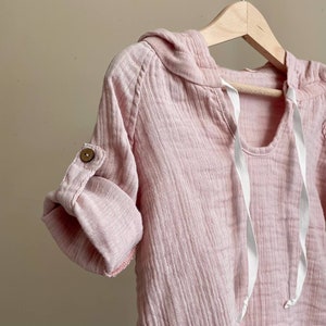 May include: A pink hooded shirt with a white drawstring and a button on the sleeve.