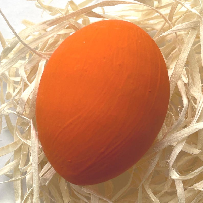 May include: A bright orange egg sits nestled in straw. The egg's smooth surface contrasts with the textured straw, creating a visually appealing composition. The egg is a solid, bright orange colour.