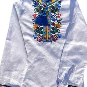 May include: White long-sleeved shirt with colorful embroidered floral pattern and blue tassel on the front. The shirt has a blue and black trim around the collar and cuffs.