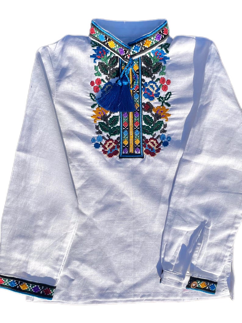 May include: White long-sleeved shirt with colorful embroidered floral pattern and blue tassel on the front. The shirt has a blue and black trim around the collar and cuffs.