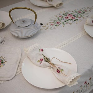 May include: A white teapot with a gold handle sits on a white linen tablecloth with embroidered floral designs. A white plate with a folded linen napkin with floral embroidery is on the table.