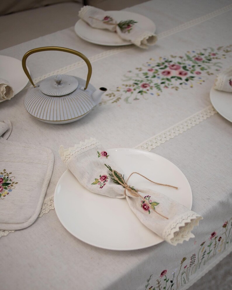 May include: A white teapot with a gold handle sits on a white linen tablecloth with embroidered floral designs. A white plate with a folded linen napkin with floral embroidery is on the table.