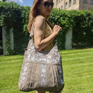 K&ouml;nnte beinhalten: Eine gro&szlig;e, taupefarbene Schultertasche mit wei&szlig;en Blumenstickereien. Die Tasche hat ein plissiertes Design und einen breiten Riemen. Das Bild wurde im Freien an einem sonnigen Tag aufgenommen. Die Person tr&auml;gt eine Sonnenbrille und ein gebl&uuml;mtes Oberteil.