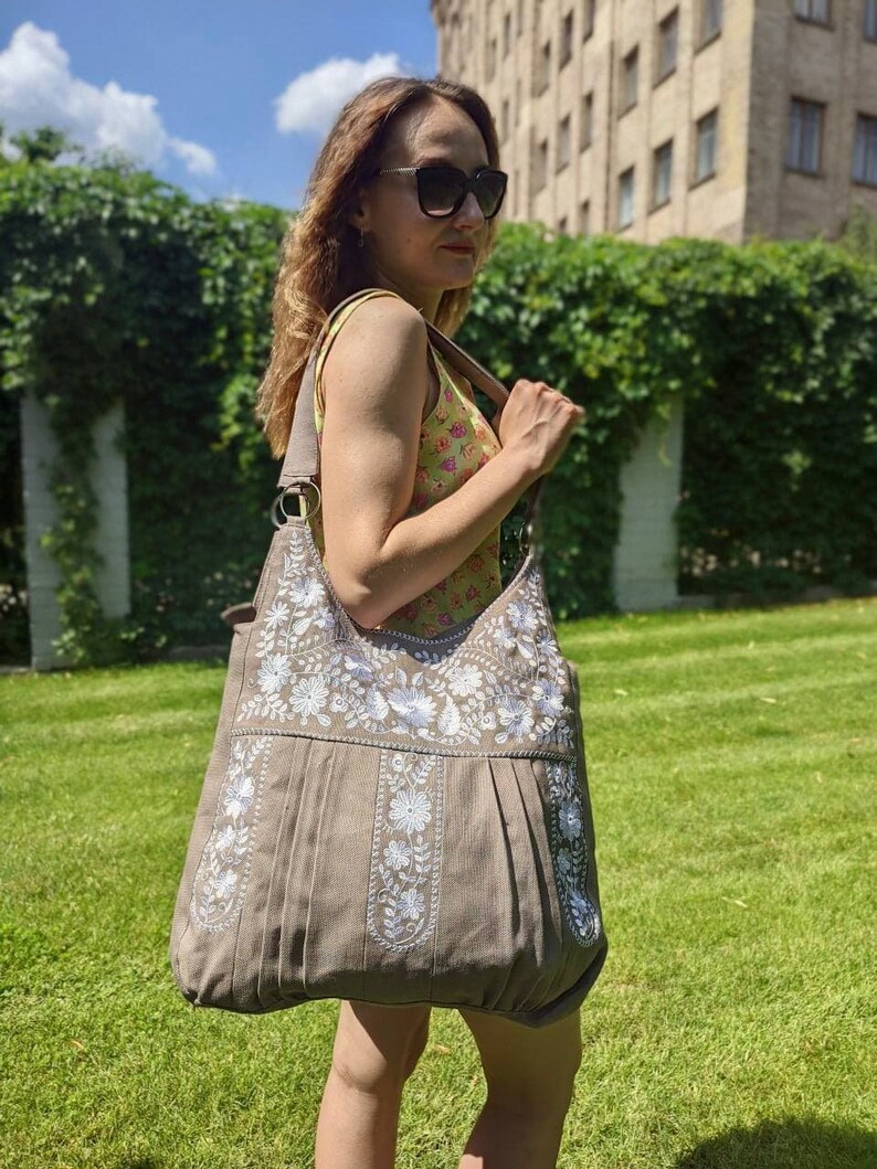 K&ouml;nnte beinhalten: Eine gro&szlig;e, taupefarbene Schultertasche mit wei&szlig;en Blumenstickereien. Die Tasche hat ein plissiertes Design und einen breiten Riemen. Das Bild wurde im Freien an einem sonnigen Tag aufgenommen. Die Person tr&auml;gt eine Sonnenbrille und ein gebl&uuml;mtes Oberteil.