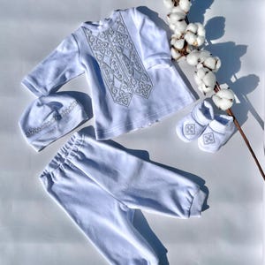 K&ouml;nnte beinhalten: Ein wei&szlig;es Baby-Outfit-Set, bestehend aus einem lang&auml;rmeligen Hemd, einer Hose, einer M&uuml;tze und Babyschuhen. Das Hemd und die M&uuml;tze haben silberne Stickereien. Das Set ist aus weichem Stoff und f&uuml;r Neugeborene konzipiert. Ein Zweig Baumwolle sorgt f&uuml;r eine dekorative Note.