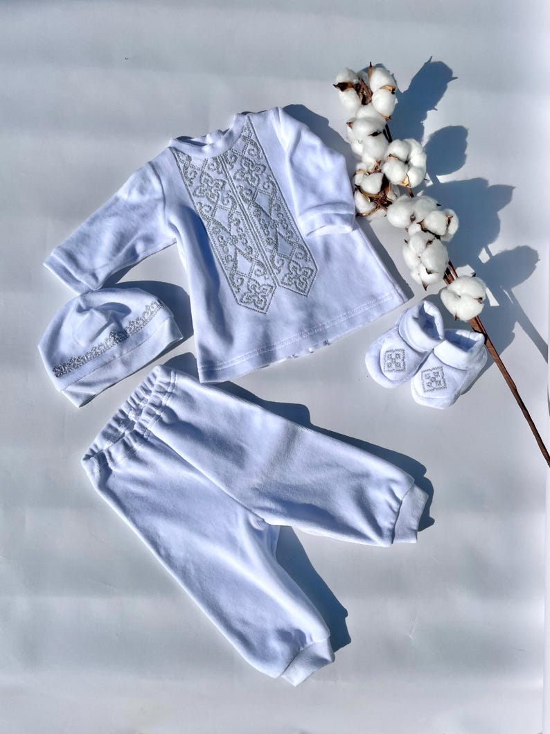 K&ouml;nnte beinhalten: Ein wei&szlig;es Baby-Outfit-Set, bestehend aus einem lang&auml;rmeligen Hemd, einer Hose, einer M&uuml;tze und Babyschuhen. Das Hemd und die M&uuml;tze haben silberne Stickereien. Das Set ist aus weichem Stoff und f&uuml;r Neugeborene konzipiert. Ein Zweig Baumwolle sorgt f&uuml;r eine dekorative Note.