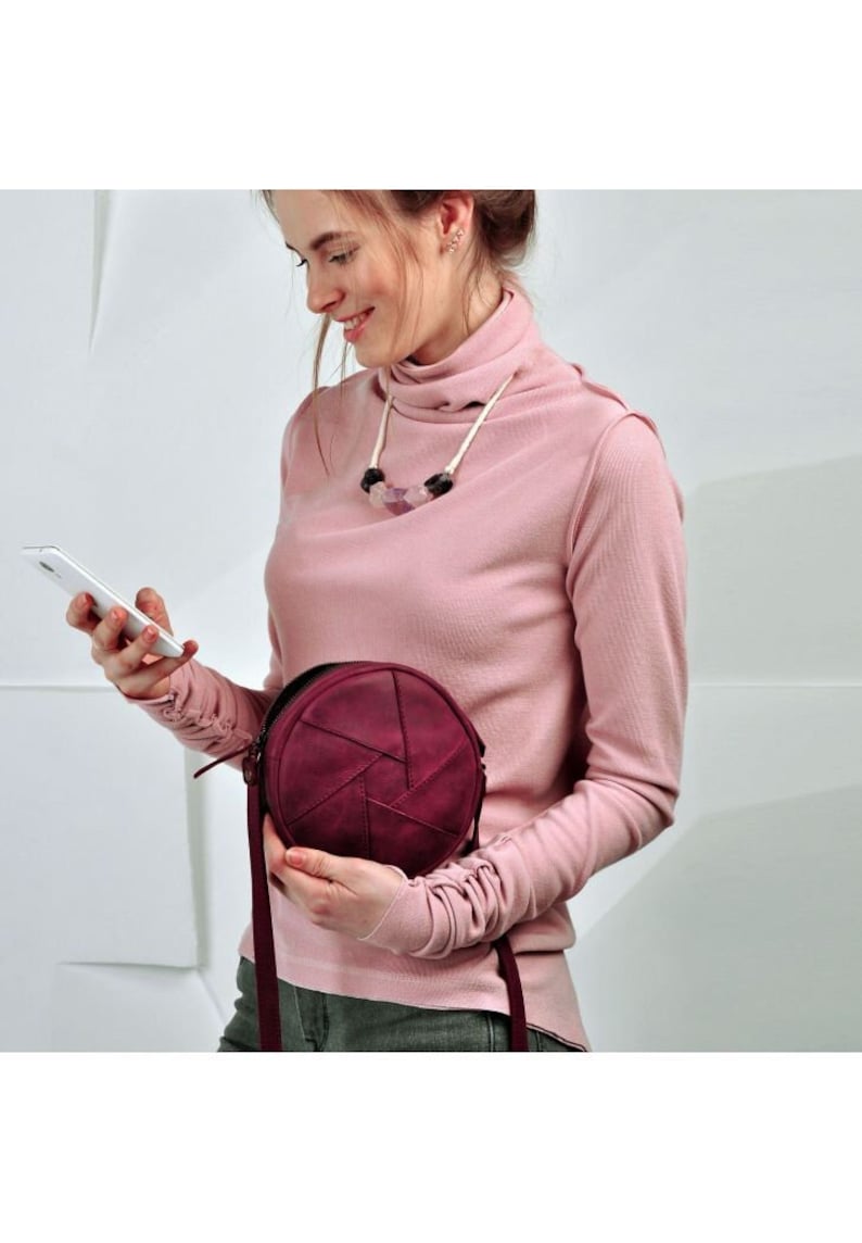 May include: A burgundy leather round handbag with a geometric design. The bag has a long strap and a zipper closure. The woman is wearing a pink turtleneck and holding a smartphone.
