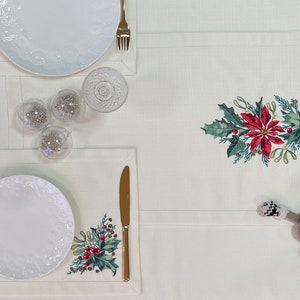 May include: A white tablecloth with a red and green embroidered poinsettia design. Two white plates with gold trim are set on placemats with matching embroidery. There are three clear glass ornaments and two gold utensils on the table.