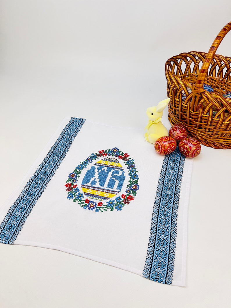 Peut inclure: Serviette de cuisine blanche brod&eacute;e avec un motif d'&oelig;uf de P&acirc;ques bleu et jaune entour&eacute; de fleurs. La serviette a des bordures bleues et blanches.
