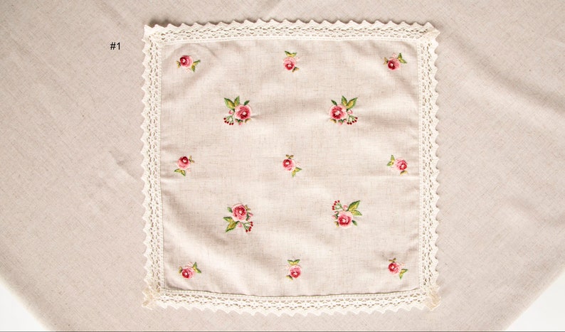 May include: Square, beige linen handkerchief with embroidered pink roses and green leaves. The handkerchief has a white lace trim around the edges. The number "1" is in the upper left corner.